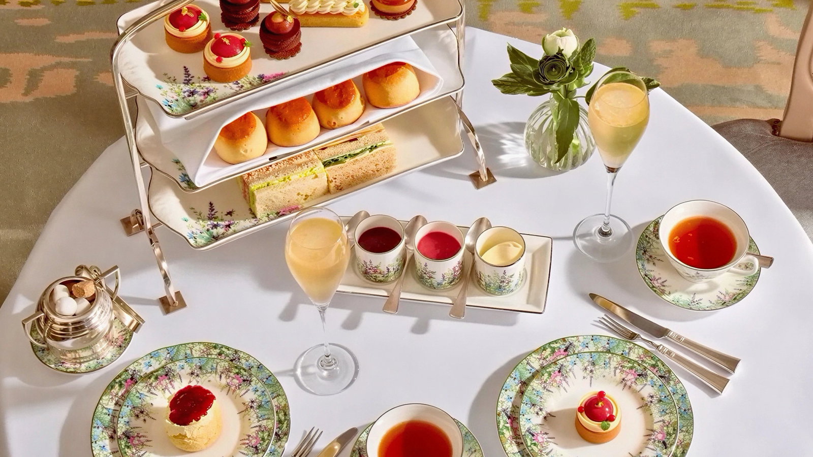 Elegant tea set on a white table with floral china, pastries, tea, a vase with a flower, and champagne glasses, creating a refined atmosphere.