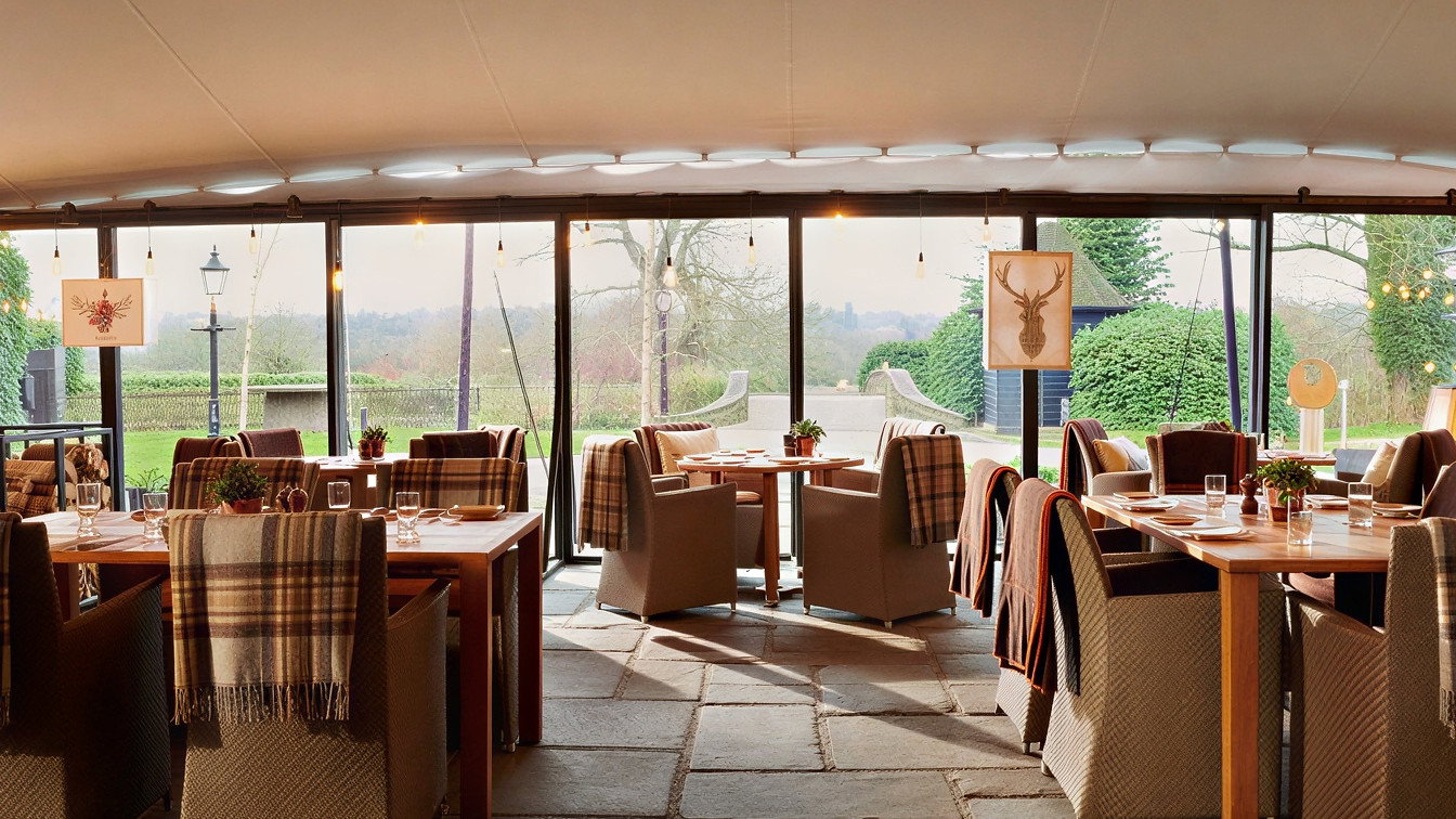 Dining area with plaid-covered chairs and wooden tables, set for a meal. Large windows show a lush garden view, creating a cozy atmosphere.