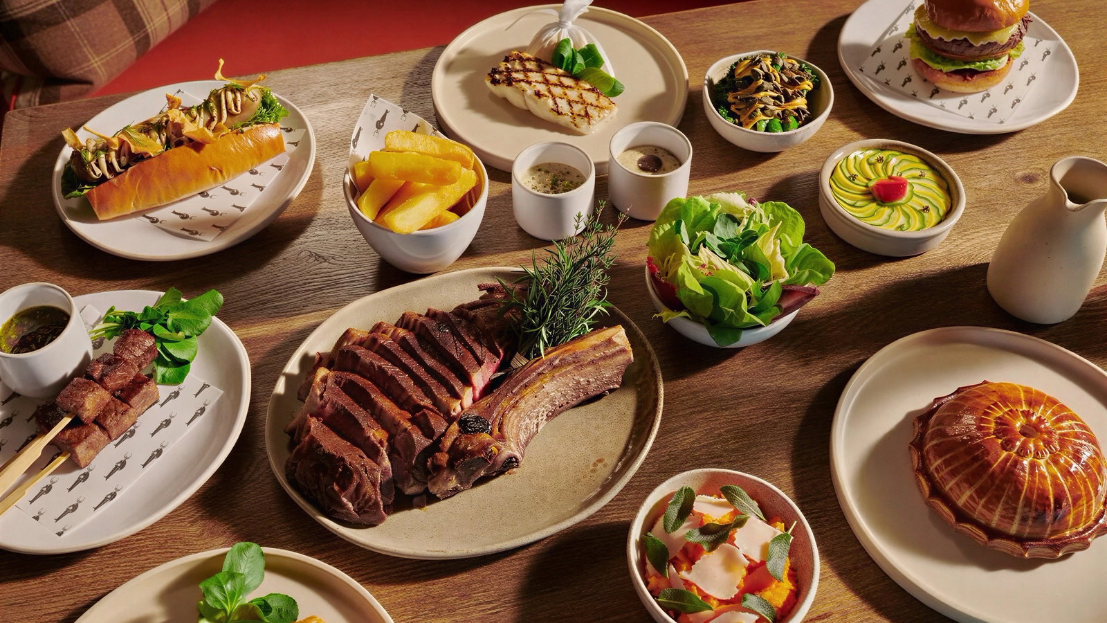 Assorted dishes on a wooden table include steak, burger, salad, fries, and desserts. Bright colors with elegant presentation.