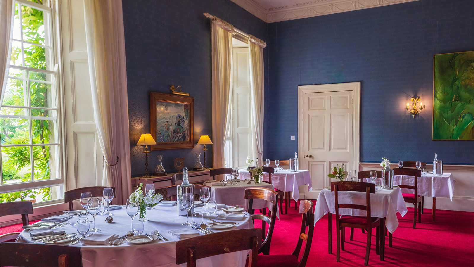 Elegant dining room with blue walls and red carpet, featuring round tables set with white cloths, glassware, and vases under natural light.