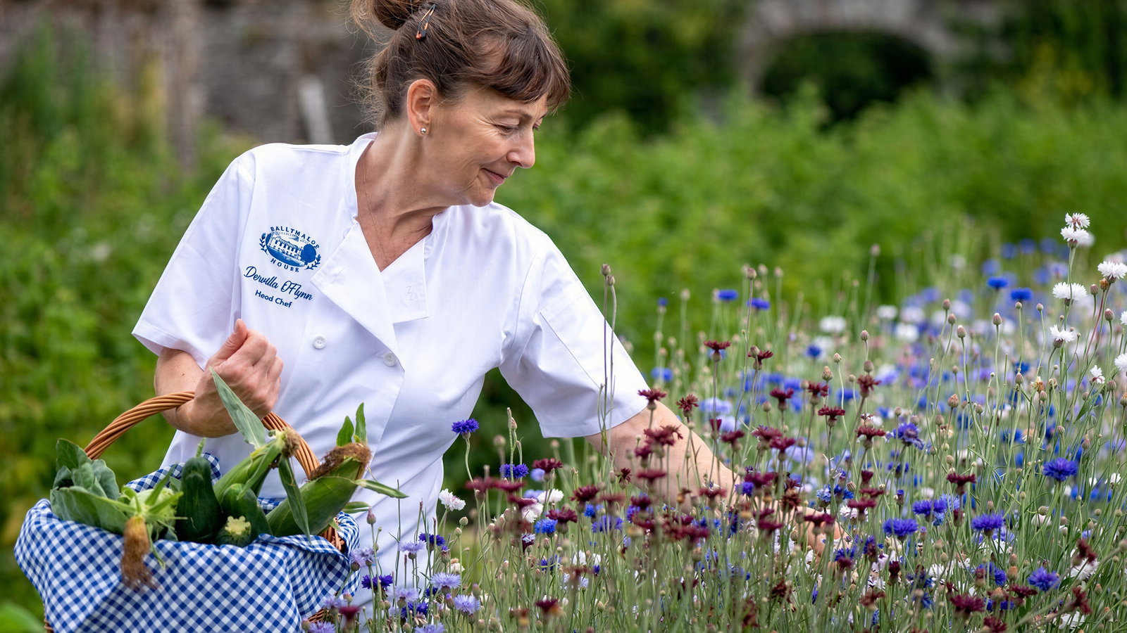 A woman in a garden picks wildflowers, carrying a basket of vegetables. She's wearing a chef's coat. Stone wall in the background.