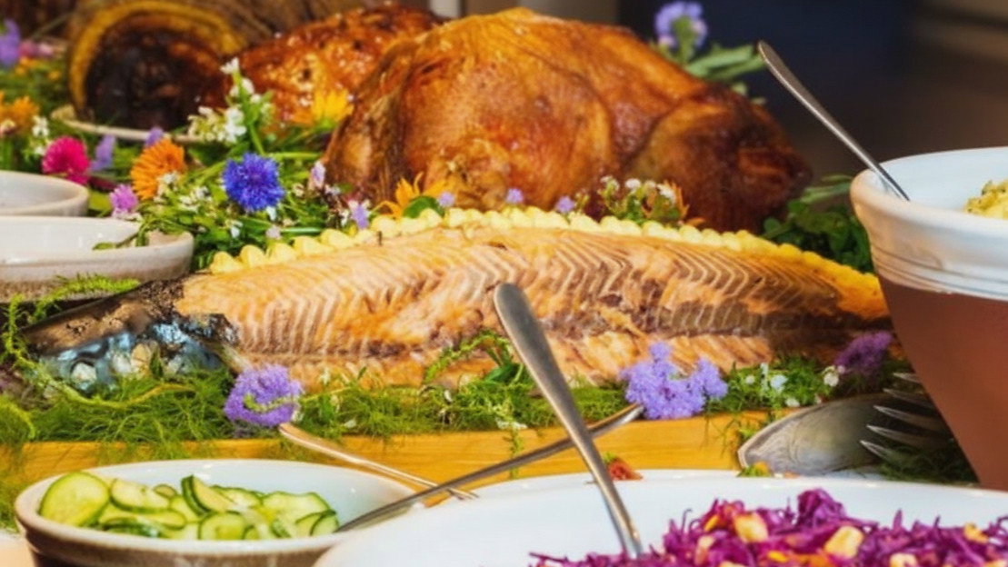 A lavish buffet with roasted meats, decorated fish, vibrant salads, and colorful flowers. Plates and utensils are arranged neatly.