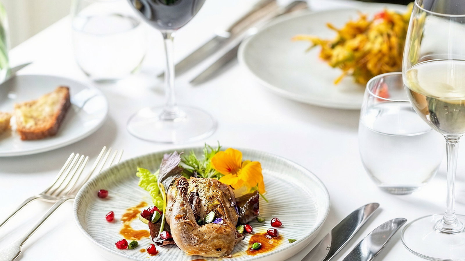 Elegant dining setup with gourmet dish, red and white wine, and water glasses on a white tablecloth. Bright, fresh ambiance.