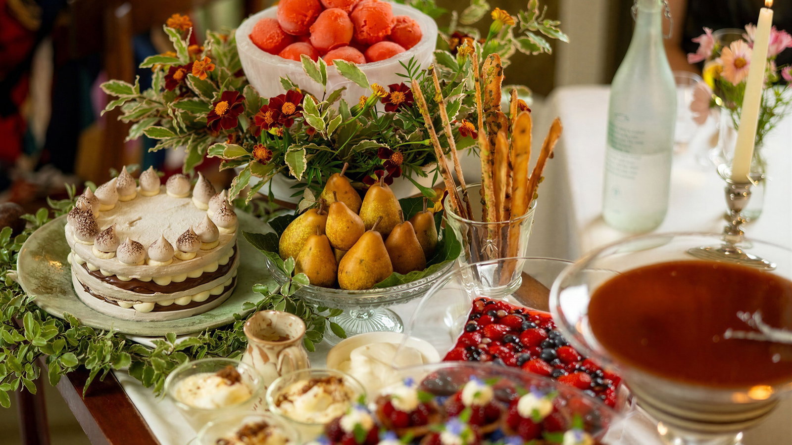 Elegant dessert table with fruit, a layered cake, pears, sorbet, and floral decor. A lit candle and glass bottle add ambiance.