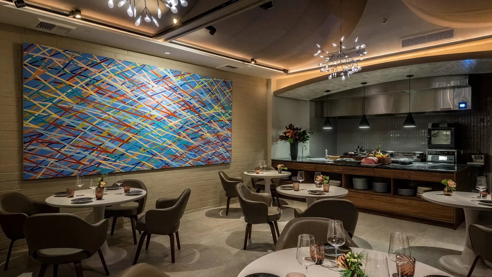 Modern restaurant with elegant tables, muted brown chairs, colorful abstract wall art, and bright overhead lighting. Kitchen area in the background.