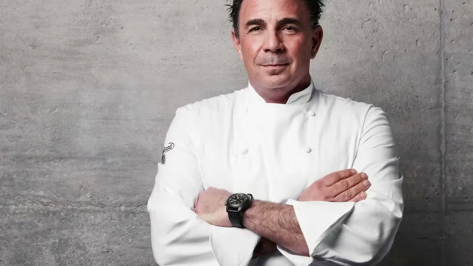 Chef in a white uniform with folded arms stands against a gray concrete wall, wearing a watch, exuding confidence and professionalism.