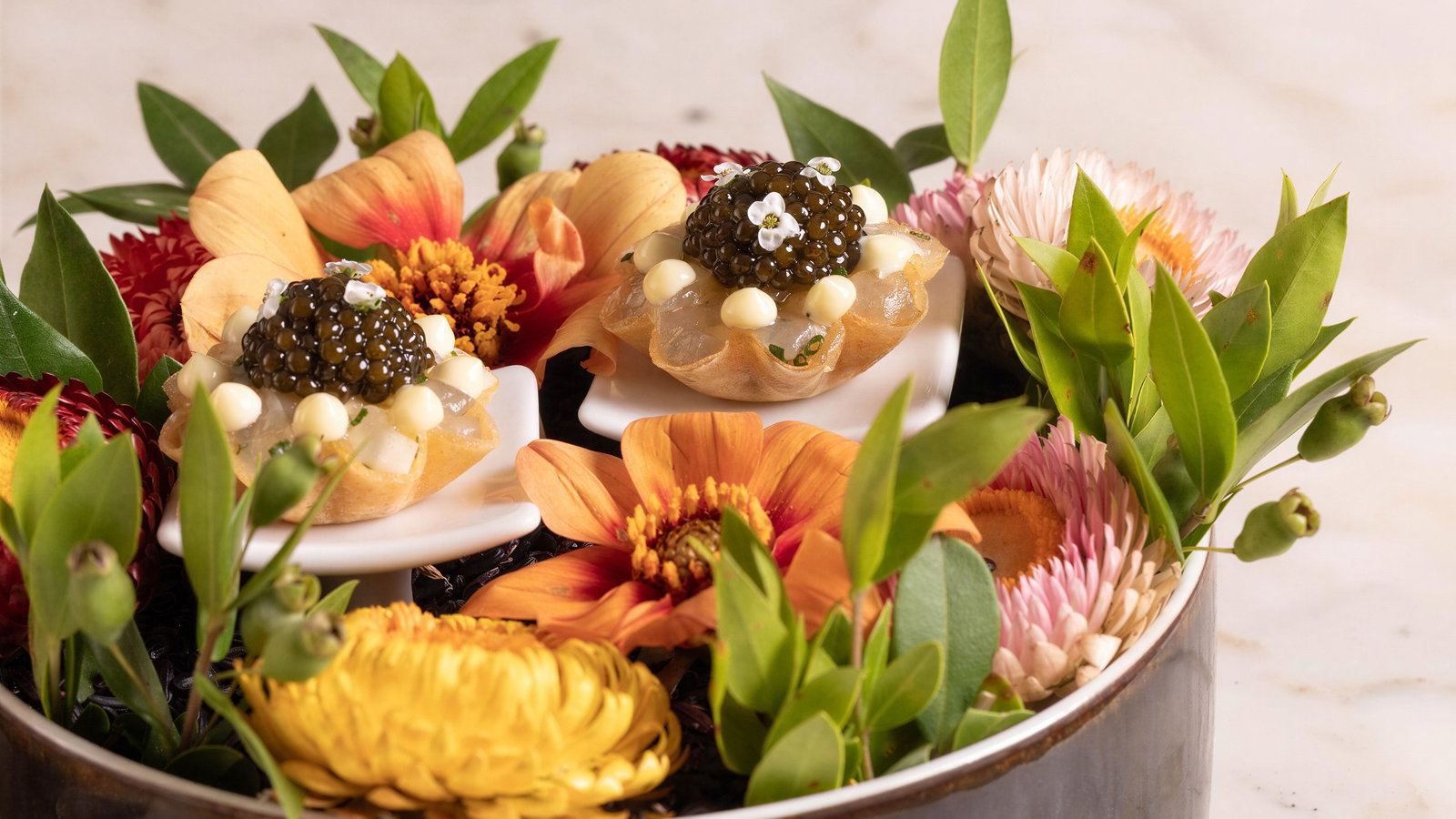 Two gourmet appetizers with caviar and cream on white dishes, surrounded by vibrant flowers and green leaves, set on a marble surface.