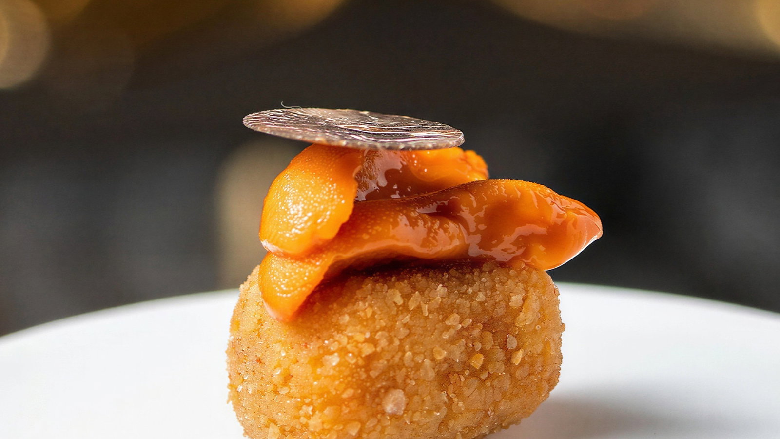 A gourmet dish features crispy, golden croquette topped with vibrant orange sea urchin and a thin, silver coin in a blurred, warm-lit setting.