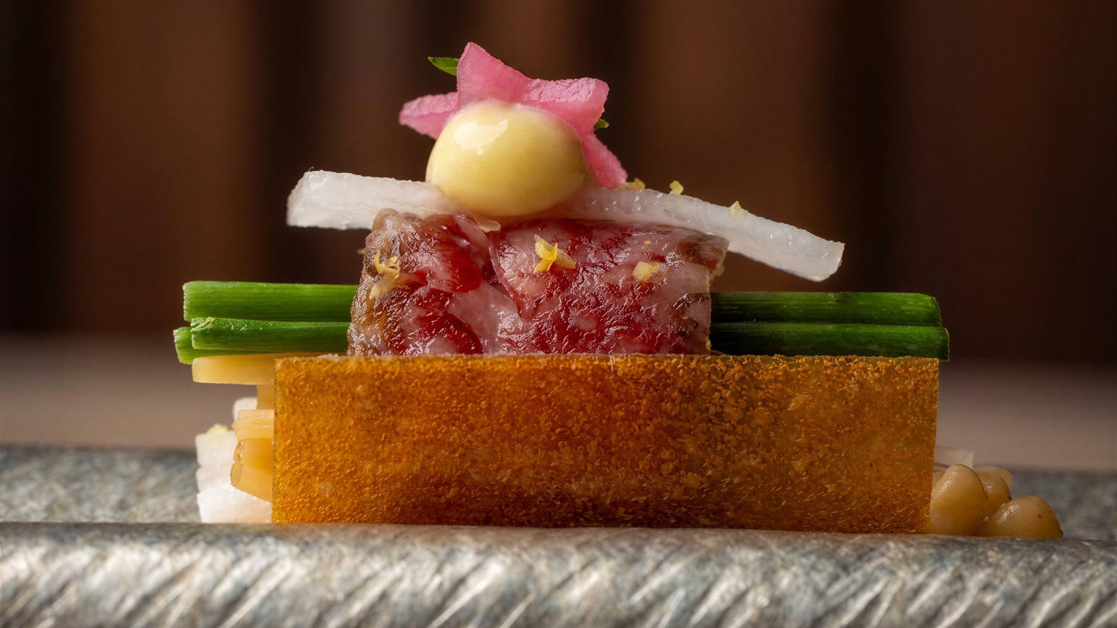 Gourmet dish with layers of meat, radish, green onions, and mustard topping. Set on a textured metallic surface, blurred background.