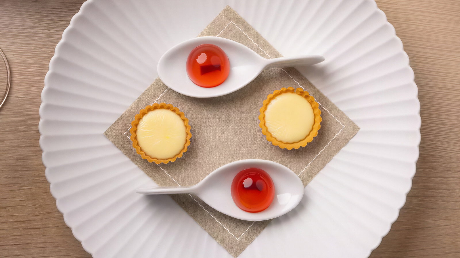 White plate with two egg tarts and red jellies in spoons on a beige napkin. Wooden table background, elegant presentation.