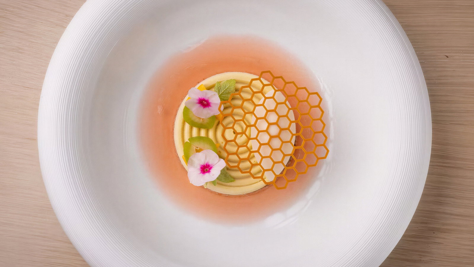 Elegant dessert with yellow honeycomb pattern, pink flowers, and green leaves on a white plate with a wood background.