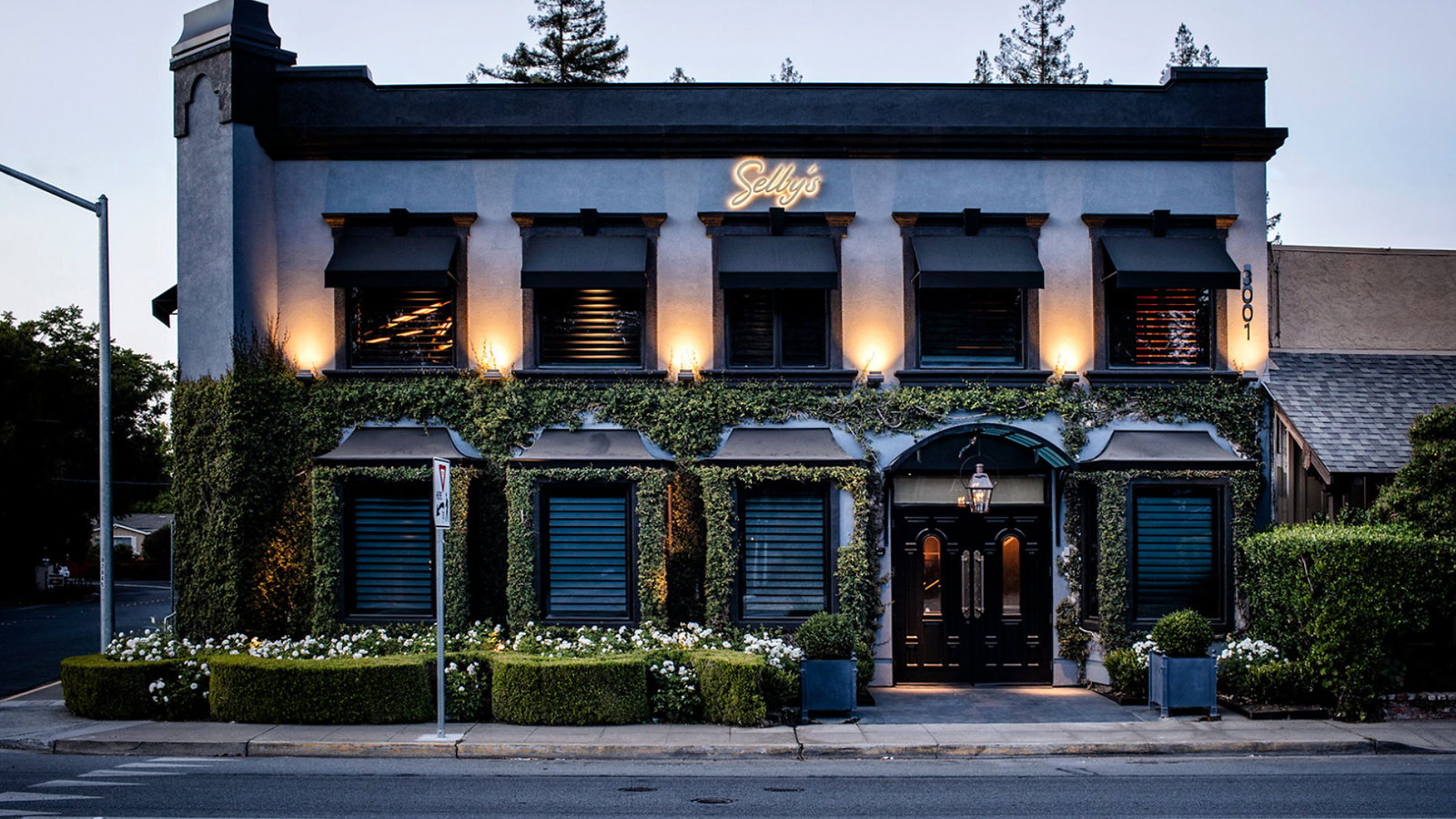 Two-story building with ivy-covered facade and large black doors. Warm lights glow above; signage reads "Sally's." Quiet street at dusk.