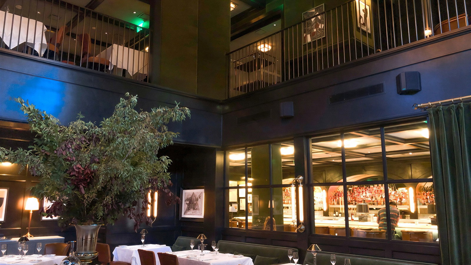 Elegant restaurant interior with white tablecloths, green walls, large plant centerpiece, and dim lighting. Empty tables and quiet ambiance.