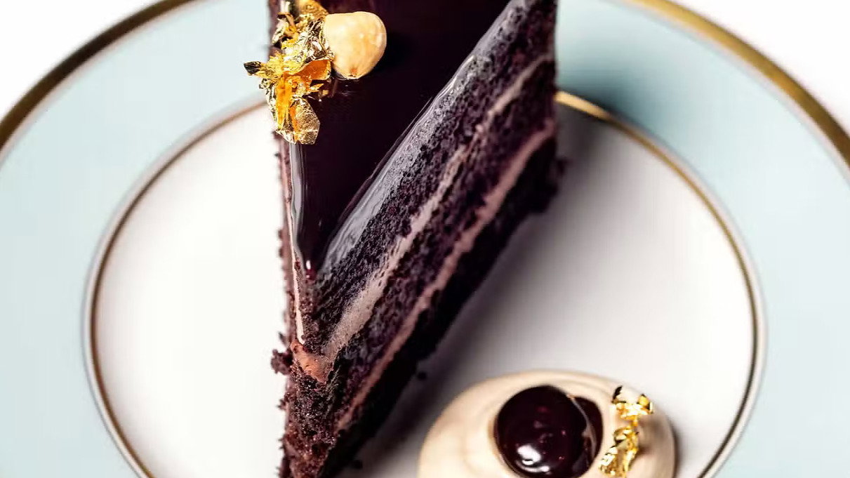 A slice of chocolate cake with gold leaf and nuts sits on a white and blue plate. Cream and chocolate garnish the side. Luxurious presentation.