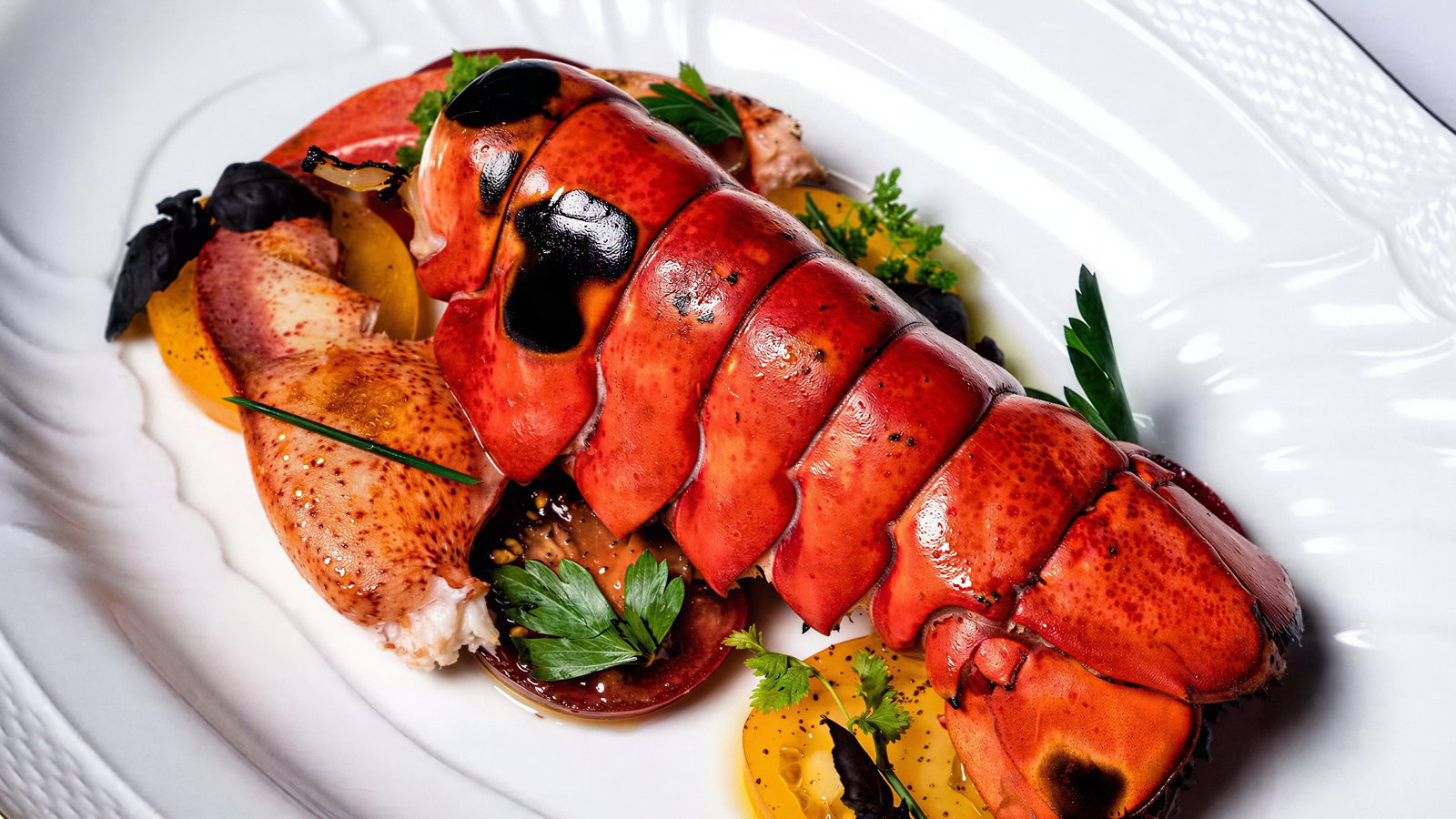 A plated lobster tail with herbs, yellow slices, and garnish on a white dish. Bright red color contrasts with fresh green leaves.