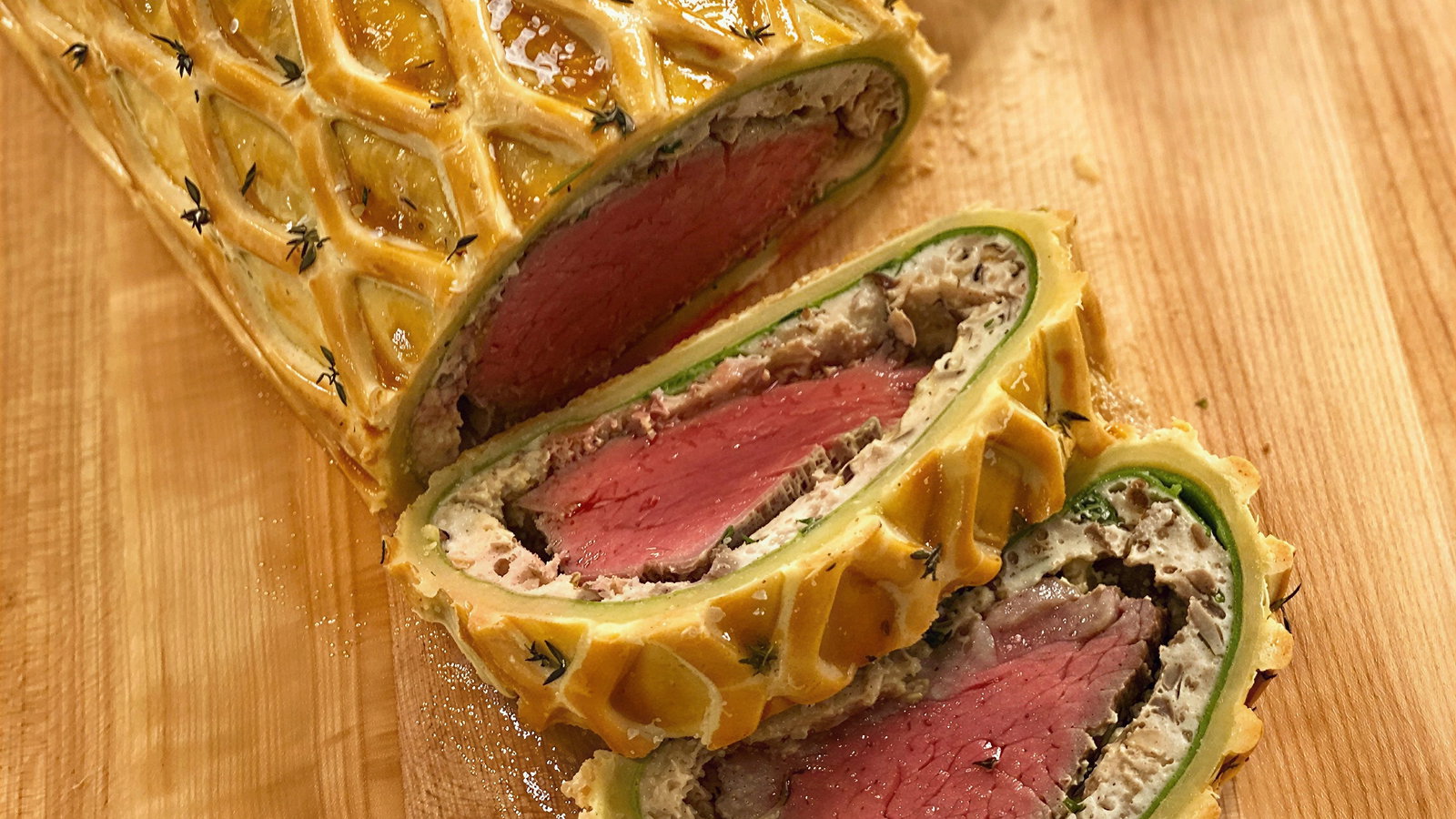Sliced beef Wellington on a wooden board, featuring a golden, lattice-pattern pastry with herbs. Juicy pink beef inside is visible.