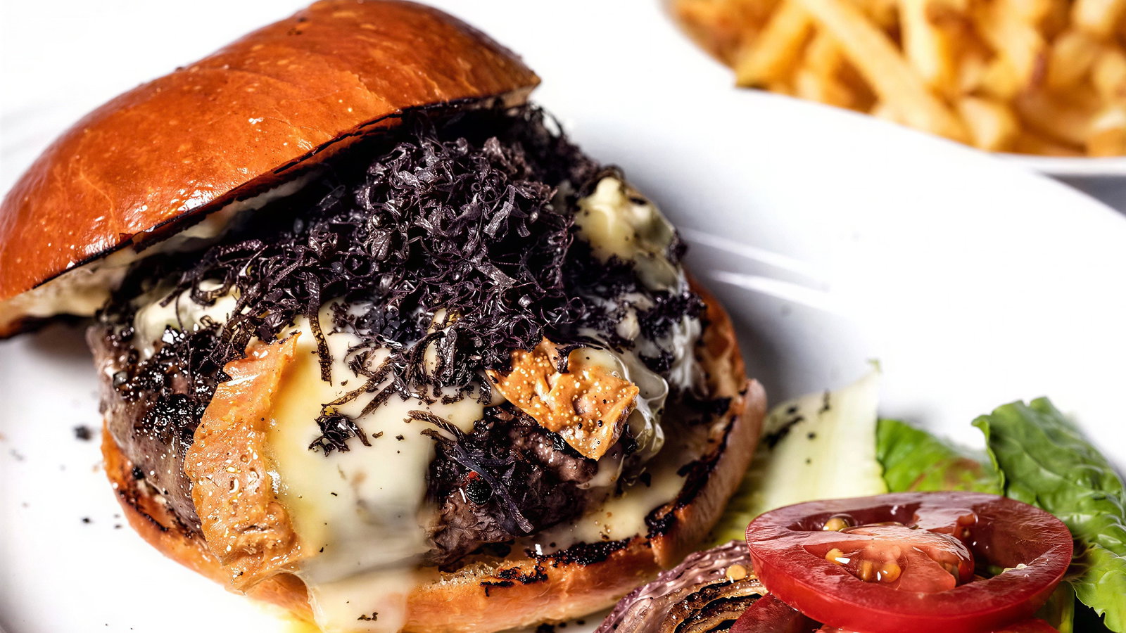 Gourmet burger with melted cheese, black truffle shavings, toasted bun. Served with lettuce, tomato, onion, and fries on a white plate.