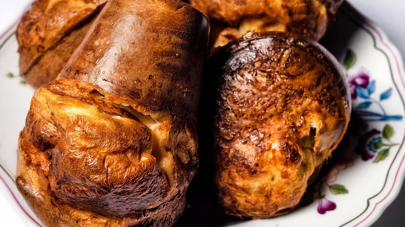 A plate of golden-brown Yorkshire puddings with crispy tops on a floral-patterned dish, set against a white background.