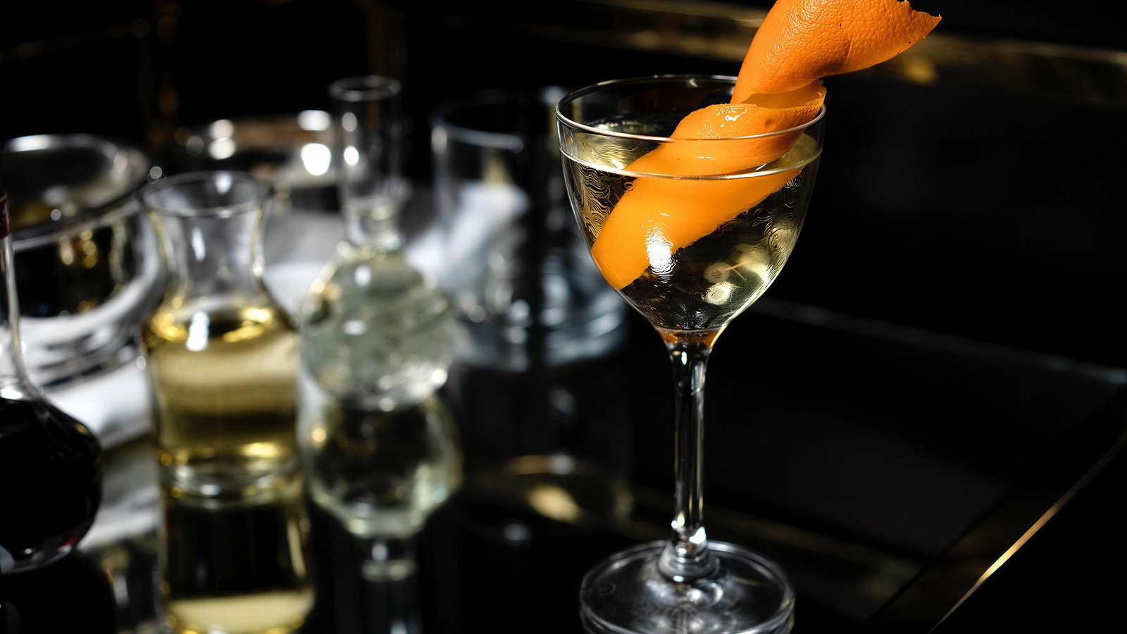 Cocktail glass with an orange peel garnish on a reflective surface, surrounded by glass bottles. Dark, elegant setting.
