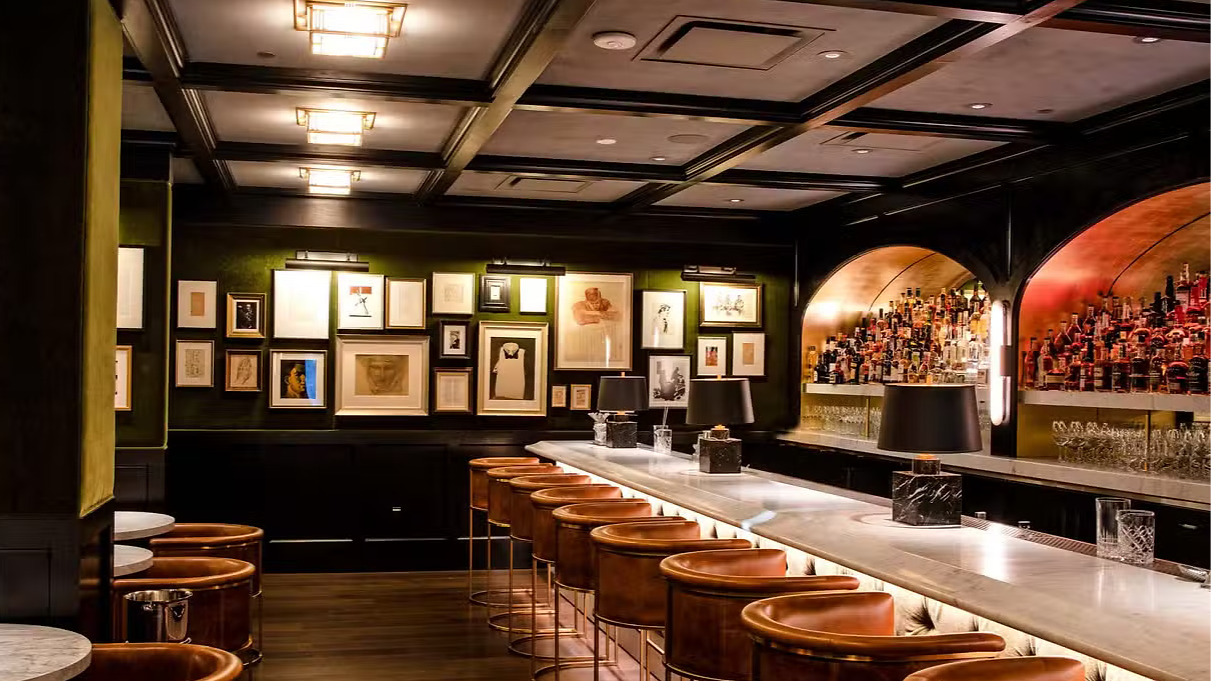 Luxurious bar with leather stools, illuminated bottles, and framed art. Warm lighting creates an inviting, elegant atmosphere.