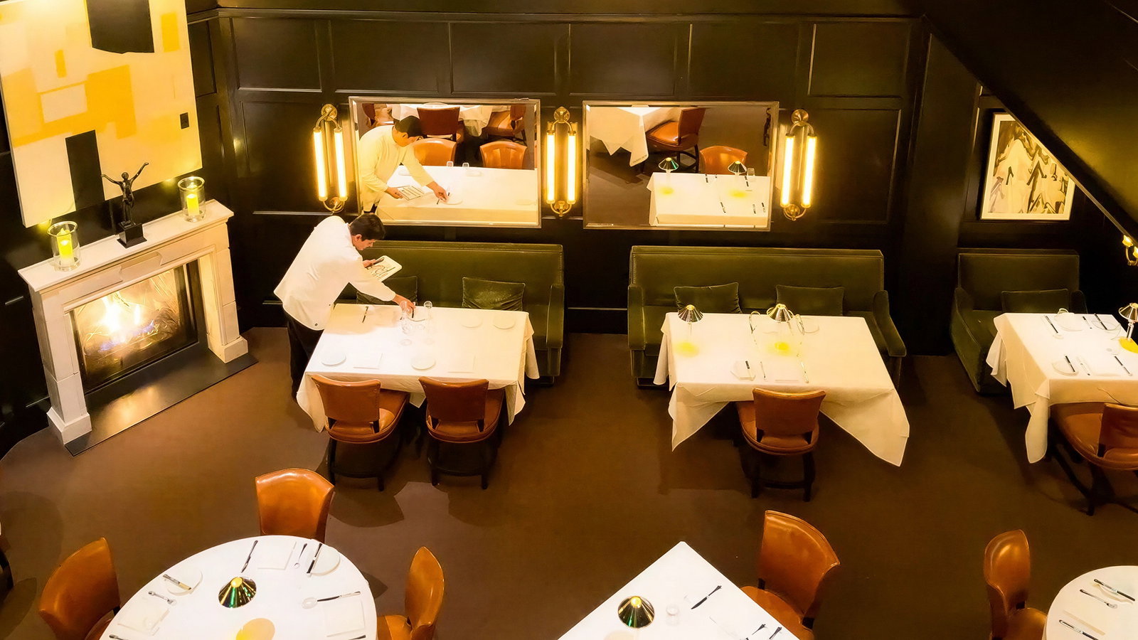 Elegant restaurant view from above. Two servers in white jackets set tables with white cloths. Warm lighting, art on dark walls, cozy ambiance.
