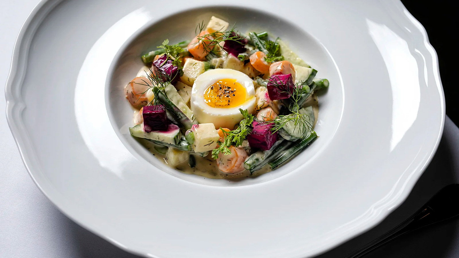 Plated salad with a soft-boiled egg, colorful vegetables, and herbs on a white dish. Elegant setting, emphasizing gourmet presentation.