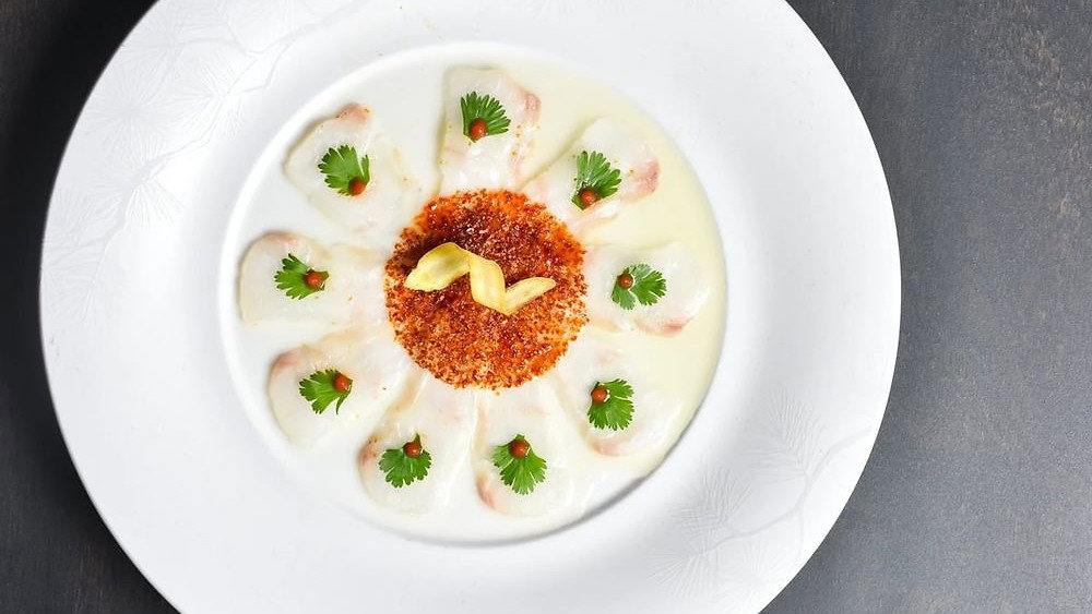 A gourmet dish on a white plate features scallops in creamy sauce, topped with herbs and spices, arranged in a floral pattern on a dark table.
