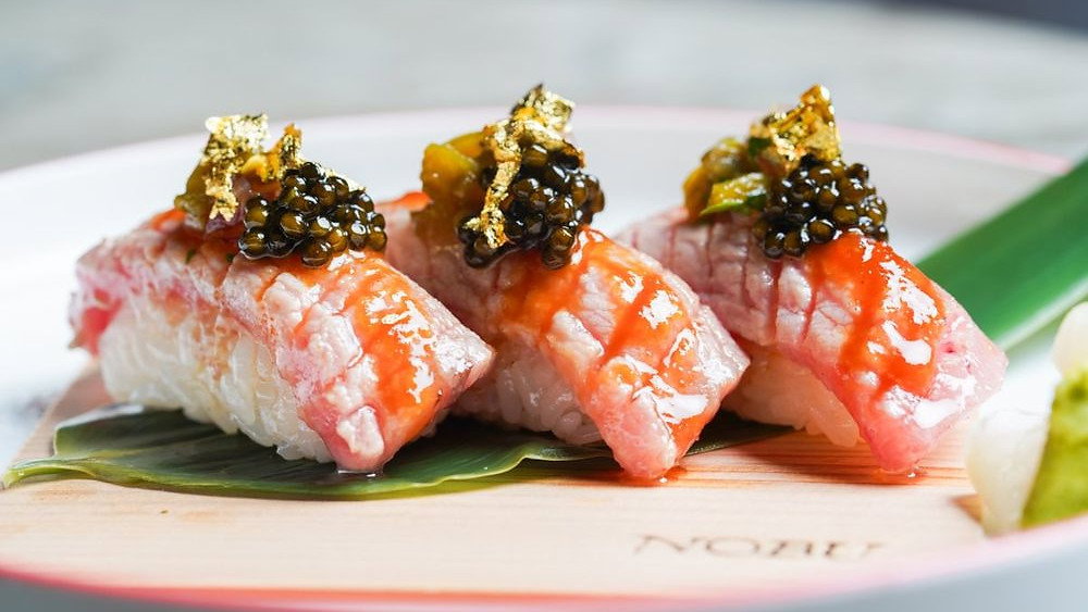 Three pieces of gourmet sushi topped with caviar and gold flakes on a green leaf. "NOBU" text is visible on the wooden board, creating a luxurious feel.