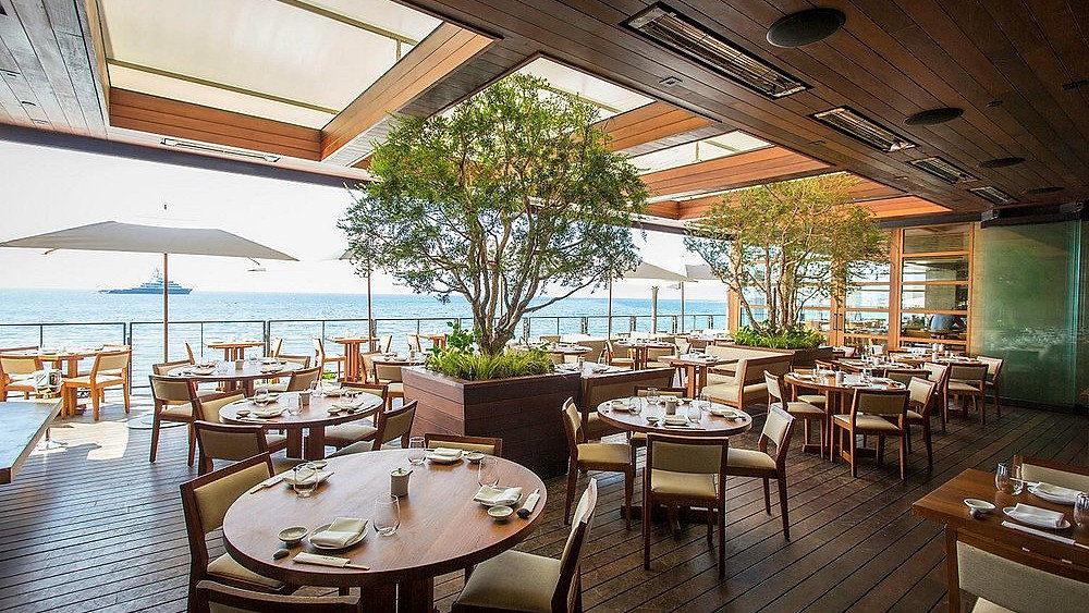 Open-air seaside restaurant with wooden tables and chairs, trees, and ocean view. Sunlit, calm atmosphere, with tables set for dining.