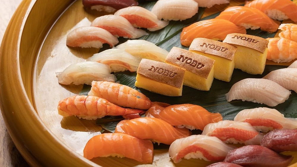 A platter of assorted sushi on a leaf-lined wooden plate. Pieces include salmon, shrimp, and tamago with "Nobu" branding. Vibrant and fresh.