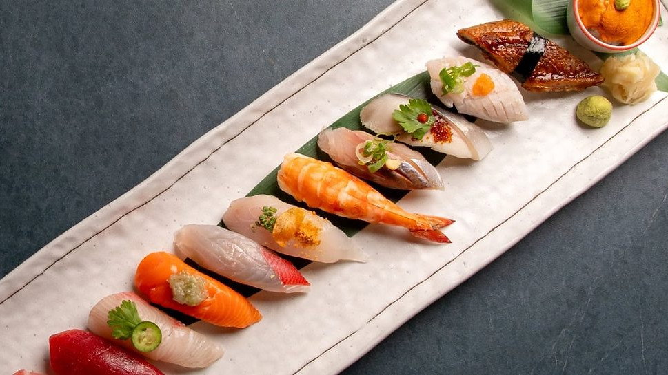 Assorted sushi on a white rectangular plate with fish, shrimp, and roe. Wasabi and ginger garnish. Gray textured background.