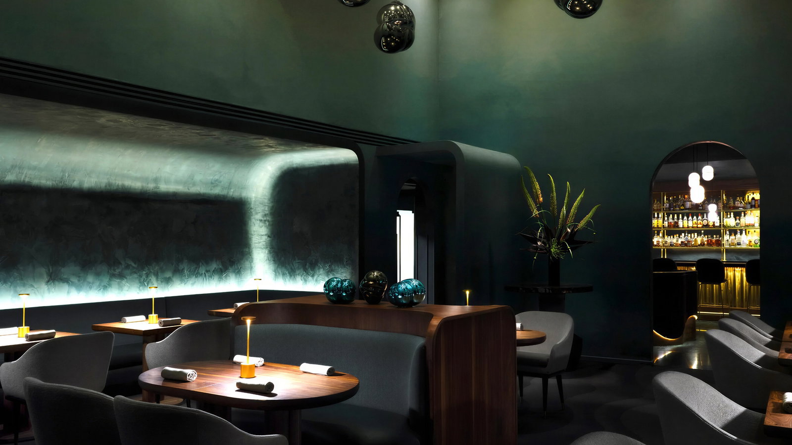 Elegant restaurant interior with dim lighting, dark teal walls, round wooden tables, and gray chairs. Bar with bottles visible through archway.