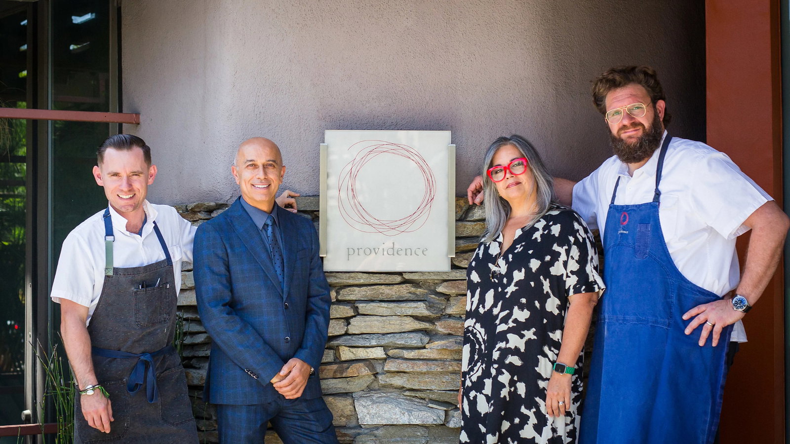 Four people stand smiling outside a building with a "providence" sign. Two in chef attire, one in a suit, and one in a patterned dress.