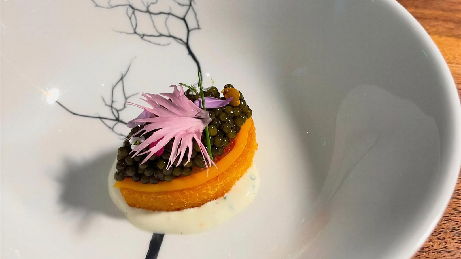 Gourmet dish on a white plate features caviar on an orange slice with a pink flower. Plate has a branch pattern, on a wooden table.