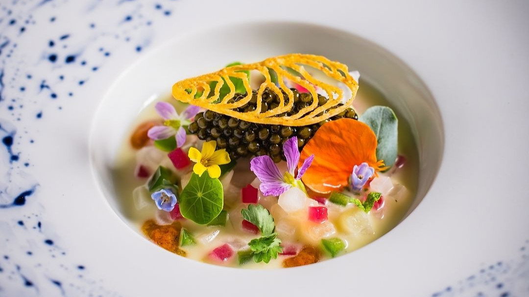 Gourmet dish on white plate, featuring vibrant edible flowers, caviar, and intricate garnish, set against a blue-speckled pattern.