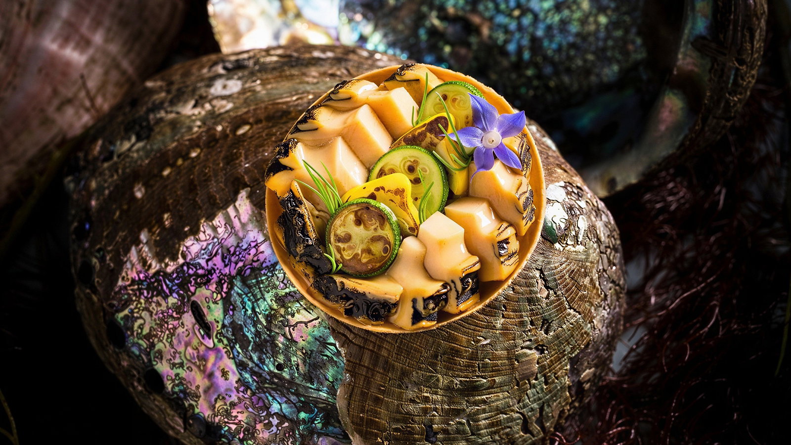 A bowl of yellow foods and a purple flower on colorful abalone shells, with dark seaweed in the background. Vibrant and elegant.