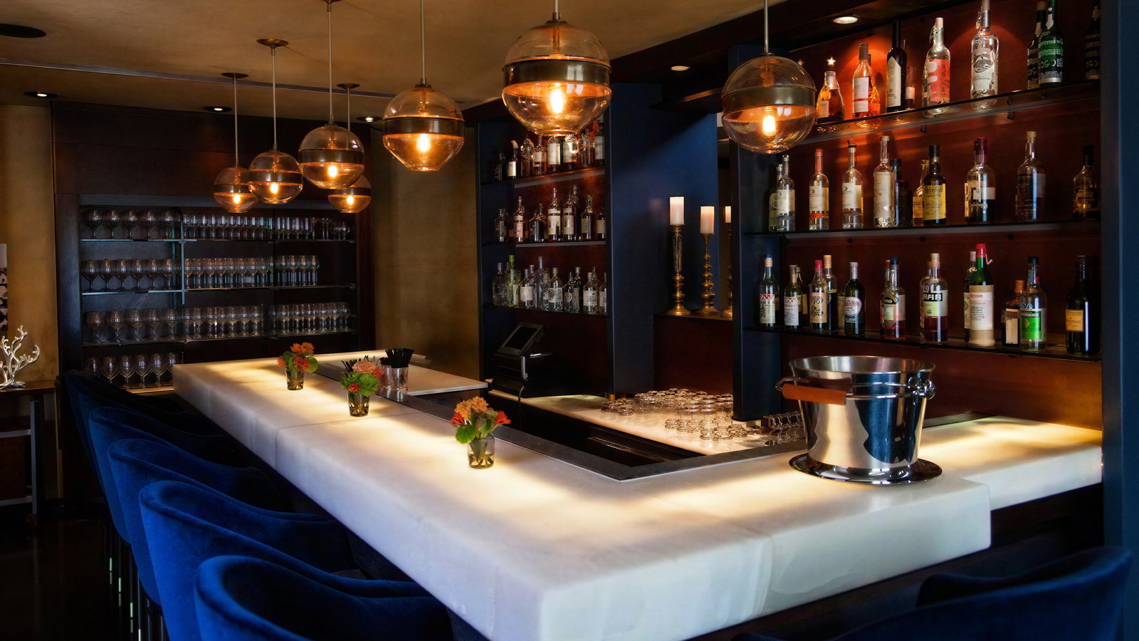 Elegant bar with glowing countertop, blue chairs, pendant lights, shelves of liquor bottles, and glasses. Warm ambiance with floral decor.