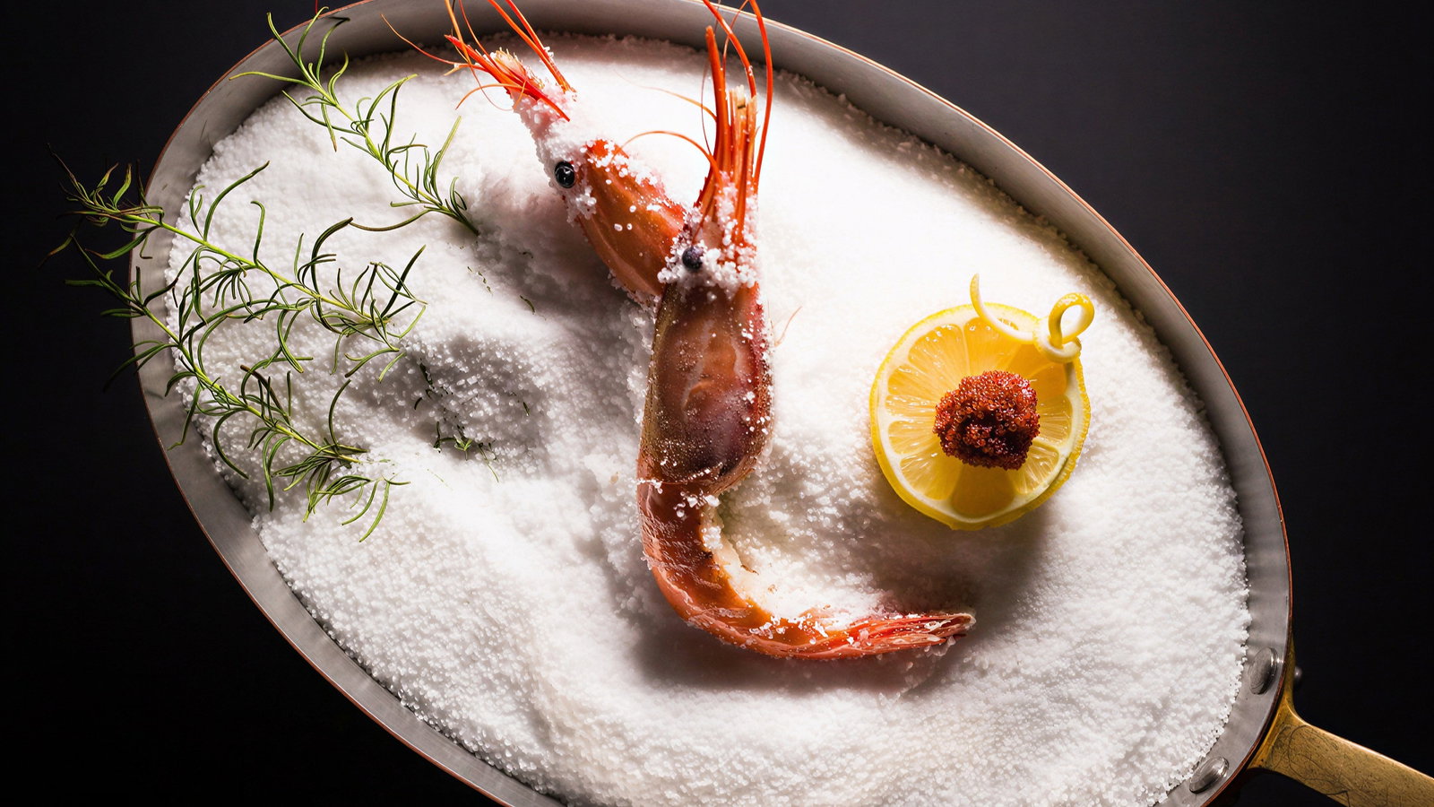 Prawn on salt bed in a pan, garnished with dill and a lemon slice with a small topping. Dark background enhances bright colors.