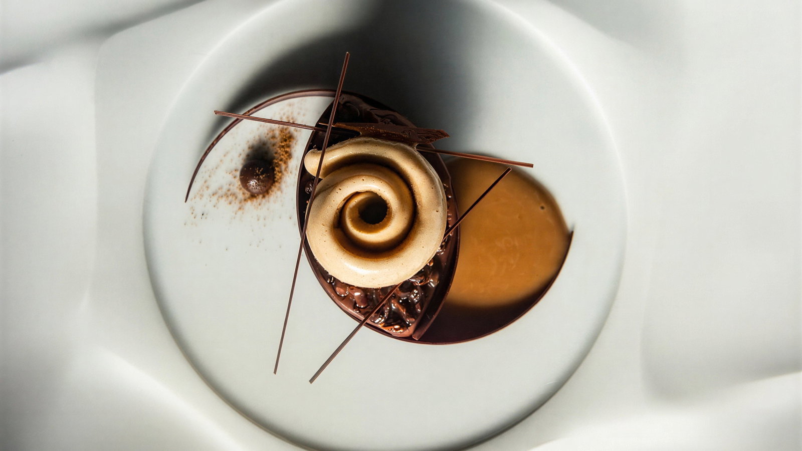 Elegant dessert with a spiral cream top, chocolate flakes, and sauce on a white plate, creating a sophisticated and artistic presentation.