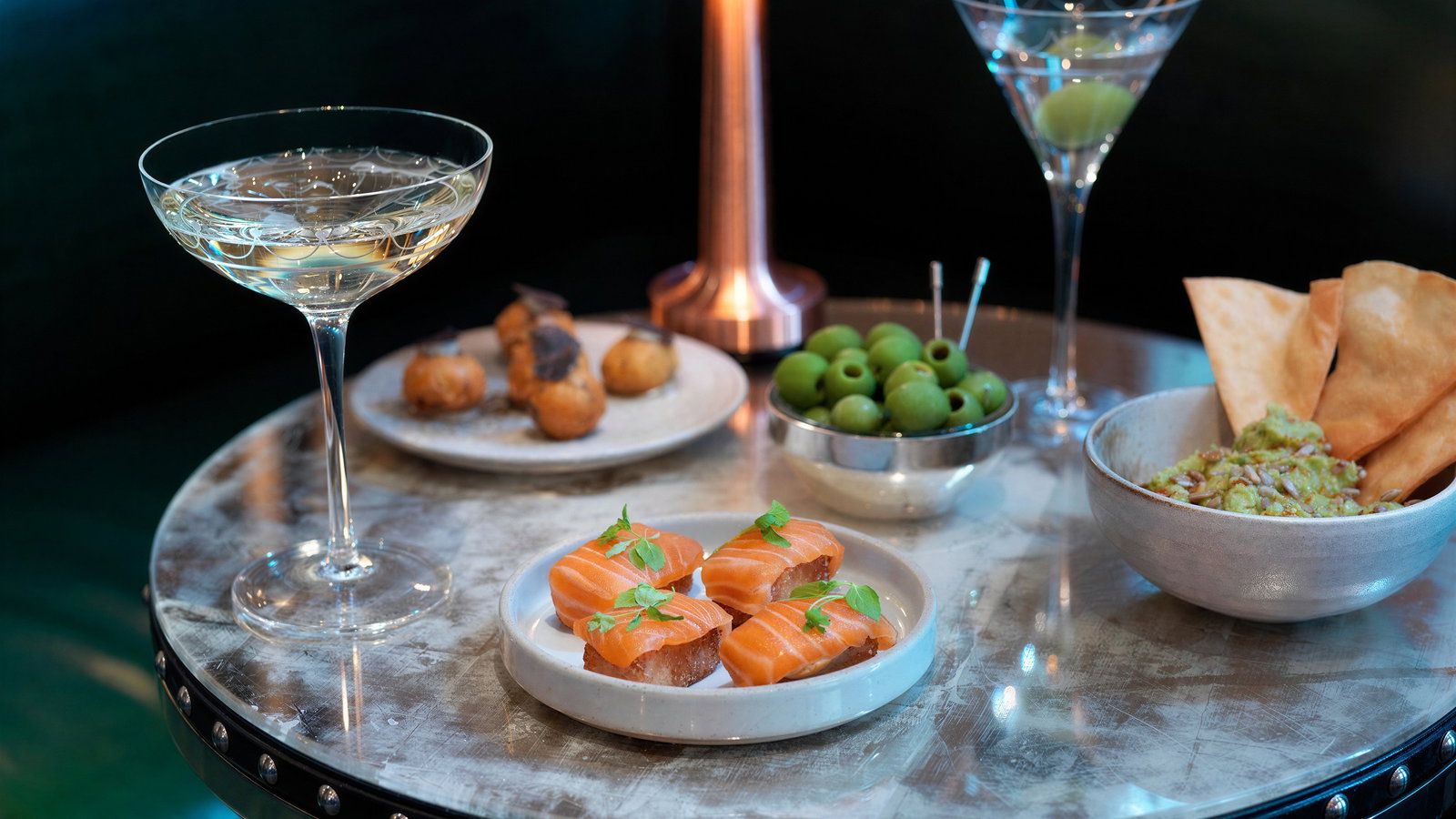 Cocktails and appetizers on a stylish round table, featuring olives, salmon canapés, guacamole, and a copper lamp in a dim setting.