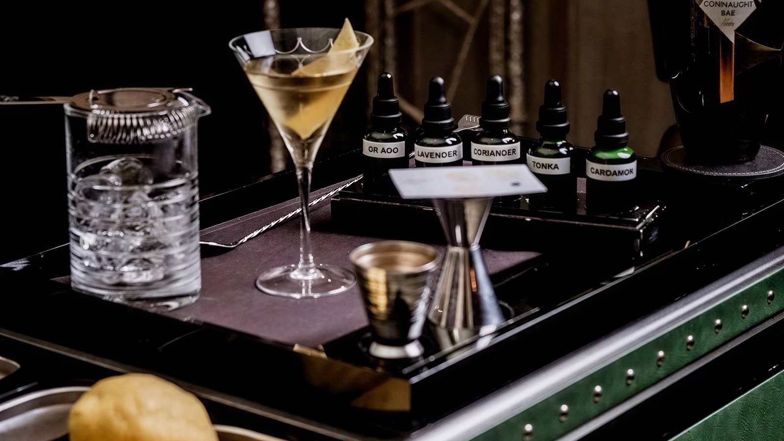 A well-lit bar counter with a martini glass, ice-filled shaker, flavor bottles labeled Lavender, Coriander, Tonka, and lemons in the foreground.