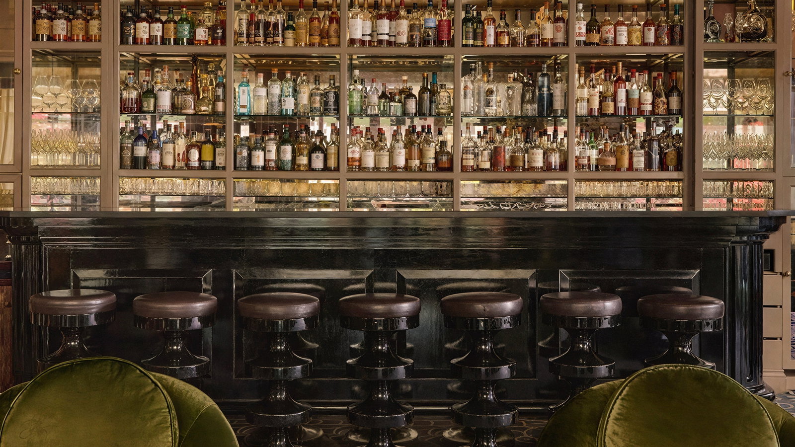 Elegant bar setting with a black counter, cushioned stools, and a backdrop of shelves filled with colorful liquor bottles, creating a cozy ambiance.