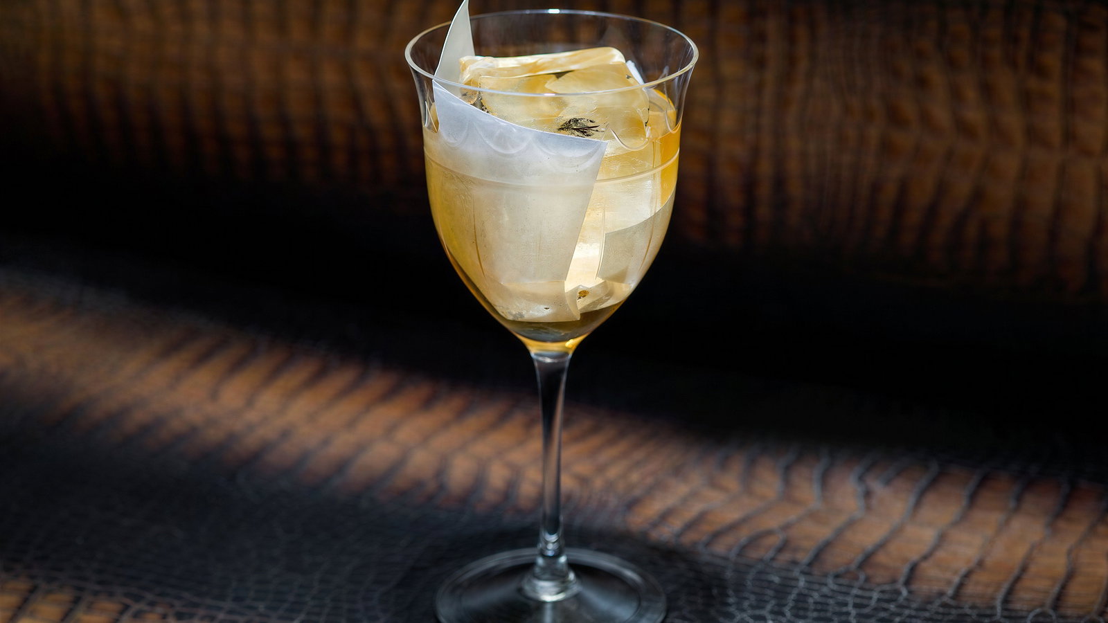 A cocktail glass with a golden drink and ice, garnished with translucent paper, sits on a textured, dark surface with a warm glow.