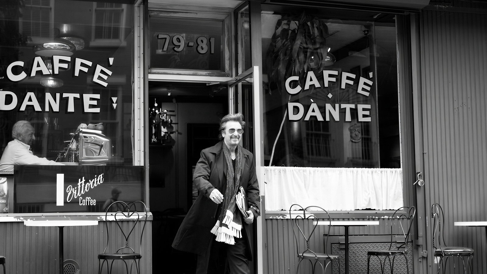 Man exits Caffè Dante, smiling in black attire, holding a scarf. Another person inside at counter. Black and white, vintage vibe.