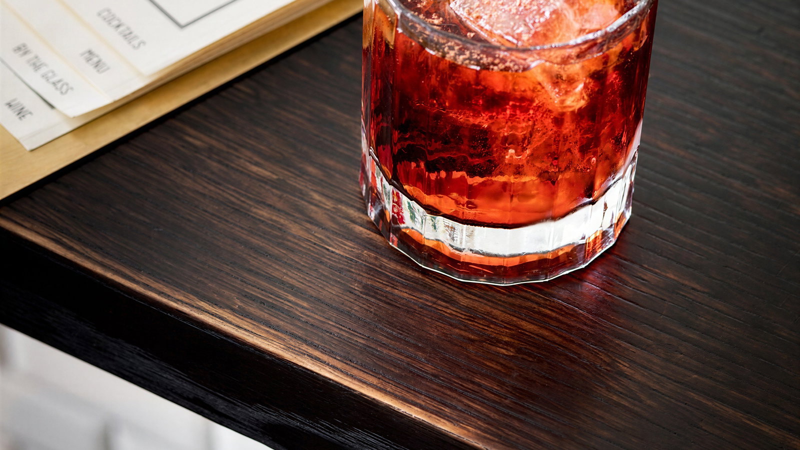 A cocktail with an orange wedge garnish and ice sits on a dark wooden table. Menus are visible in the background, creating a cozy vibe.