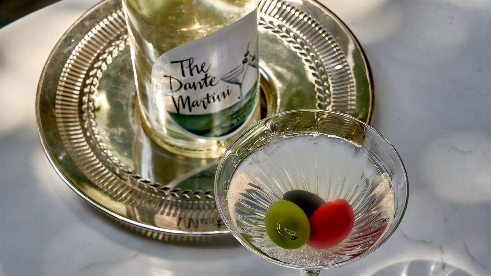 Bottle labeled "The Dante Martini" and martini glass with olives on a silver tray. Set on a white table, creating a sophisticated mood.