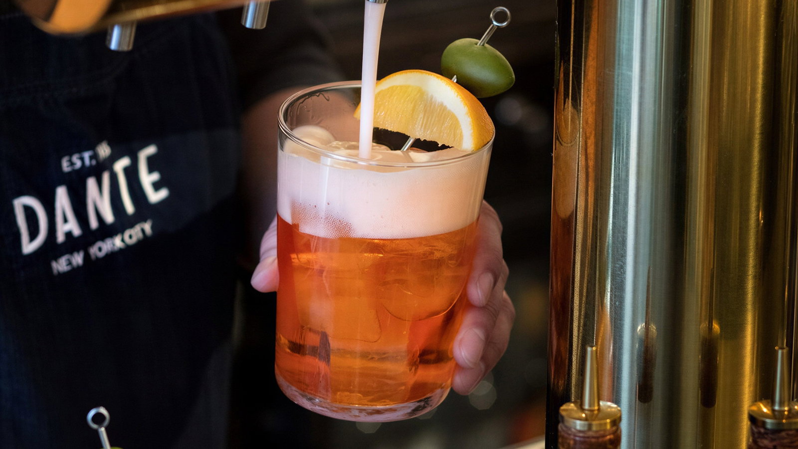 Bartender pours orange cocktail with foam from tap, garnished with orange slice and olive. Wearing "DANTE" apron. Copper tap visible.