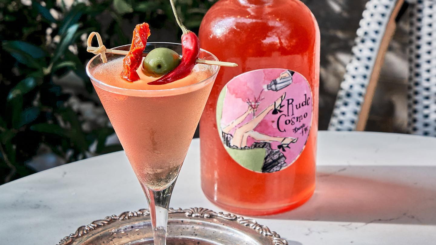 Cocktail garnished with olive and chili on silver tray with pink coaster. Reddish bottle labeled "Rude Cosmo" beside, leafy background.