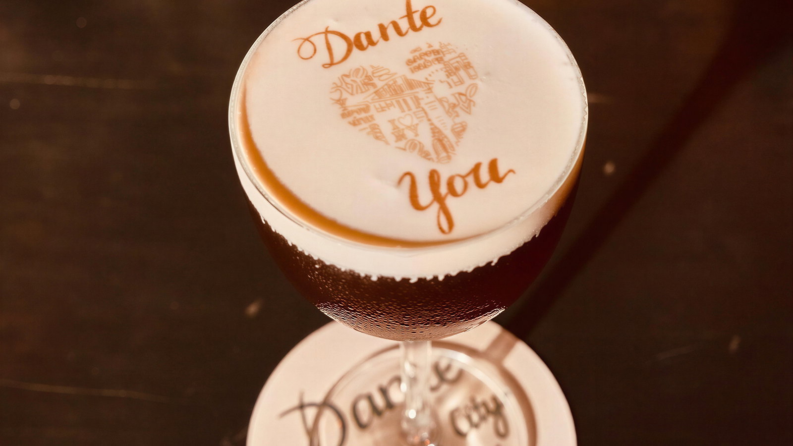 A cocktail with "Dante" and "You" in foam sits on a coaster with "Dante, New York City, Est. 1915" written, on a dark bar counter.