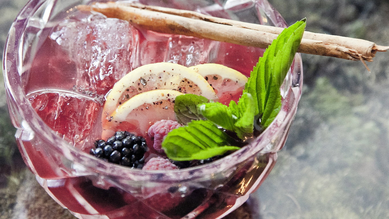 Pink cocktail in a crystal glass with ice, lemon slices, blackberries, raspberries, mint, and a cinnamon stick on a textured background.